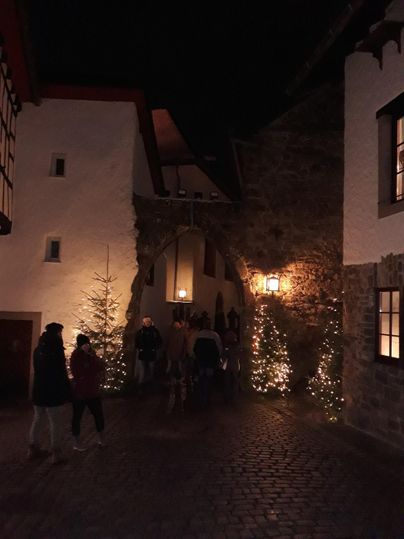Eine malerische Gasse bei Nacht, beleuchtet von warmem Licht und festlichen Dekorationen. Menschen schlendern entspannt vorbei, während ein Weihnachtsbaum im Vordergrund steht.