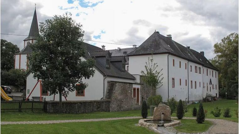 Ein historisches Gebäude mit weißen Wänden und einem grauen Dach, umgeben von Bäumen und Rasen. Im Hintergrund ist ein Kirchturm sichtbar.
