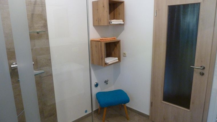A modern bathroom with a shower and a wooden shelf. A blue stool is in the corner, and the door has a window.
