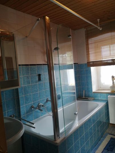 Een lichte badkamer met een bad en een douchewand. De muren zijn betegeld in een vriendelijke blauwe kleur, en er is een raam met jaloezieën.