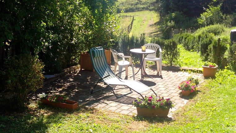 Ein einladender Gartenbereich mit einem Sonnenliegestuhl und einem weißen Stuhl. Umgeben von Blumen und einem schönen grünen Hintergrund.