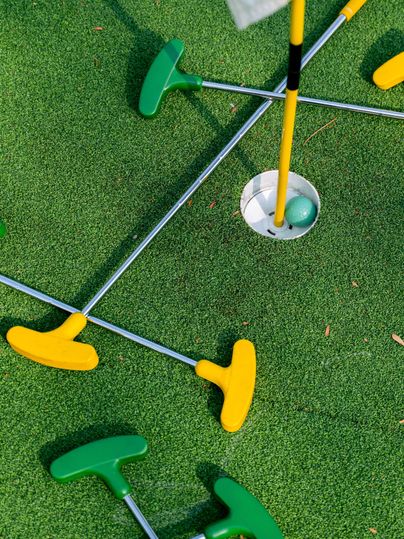 In der Mitte des Bildes sieht man ein Loch, indem ein grüner Golfball liegt. Um das Loch liegt grüner Kunstrasen, auf dem mehrere Schläger liegen. Die Schläger haben gelbe und grüne Schlägerköpfe, der Schaft ist metallfarbend.