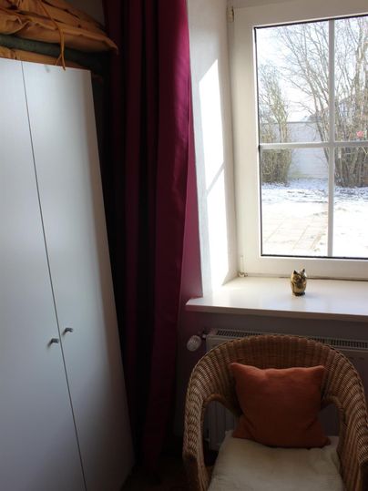 Een gezellige kamer met een rieten stoel, een kussen en een witte kast. Door het raam zie je een winterse buitenruimte.
