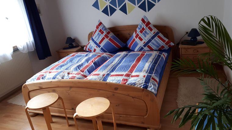 Ein gemütliches Schlafzimmer mit einem großen Bett und bunten Kissen. Zwei kleine Hocker stehen neben dem Bett, und es gibt eine Pflanze im Raum.