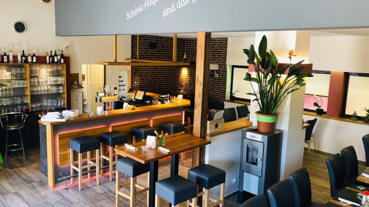Modernes Restaurant mit Bar, Holztischen und Stühlen. An der Wand steht "Schöne Augenblicke sind das Glück des Lebens". Pflanzen und Gläser dekorieren den Raum.