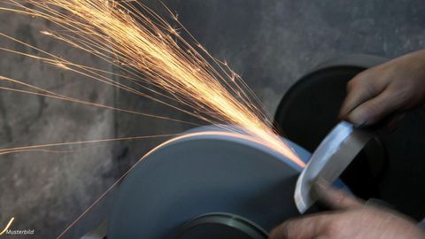 Ein Messer wird an einem Schleifstein geschärft, während Funken sprühen. Hände halten das Messer fest.