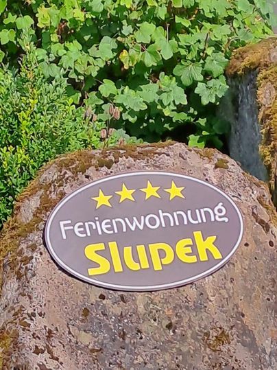Ein Schild mit dem Namen "Ferienwohnung Slupek" auf einem großen Stein. Im Hintergrund sind grüne Pflanzen sichtbar.