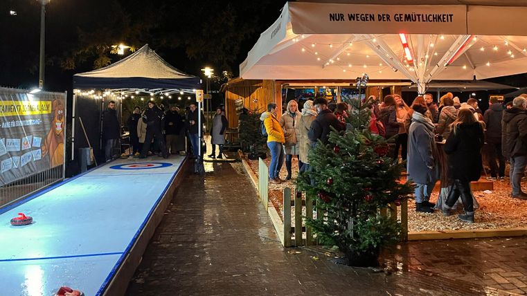 Eine festliche Winteratmosphäre mit vielen Menschen, die sich um ein überdachtes Gebiet versammeln. Auf der linken Seite sieht man eine Eisbahn mit bunten Curling-Steinen.