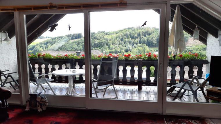 Ein gemütlicher Balkon mit Blick auf grüne Hügel und blühende Pflanzen. Auf dem Tisch stehen Stühle und ein runder Tisch, ideal zum Entspannen.