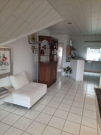 Ein heller und einladender Raum mit weißen Fliesen und einer gemütlichen Sitzgelegenheit. Im Hintergrund befindet sich eine offene Küche und ein Holzregal.