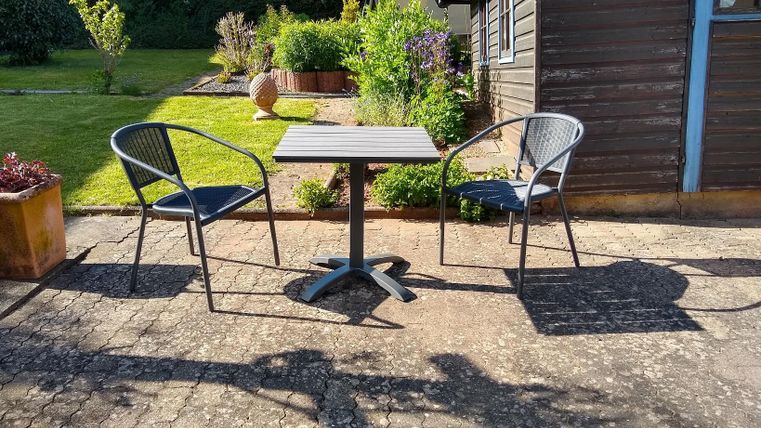 Ein kleiner Tisch mit zwei Stühlen steht auf einem gepflasterten Bereich im Garten. Im Hintergrund sind Pflanzen und ein Gebäude sichtbar.