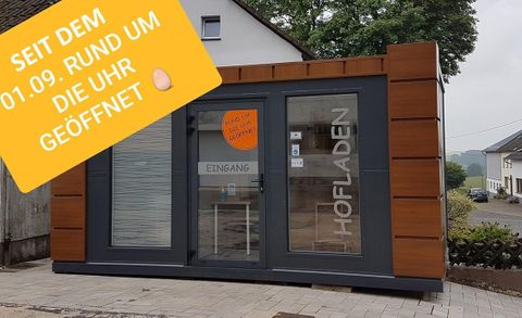 Moderne boerderijwinkel met glazen voorzijde en houten elementen. Bord: "Sinds 01.09. 24/7 geopend". Plattelandsomgeving.