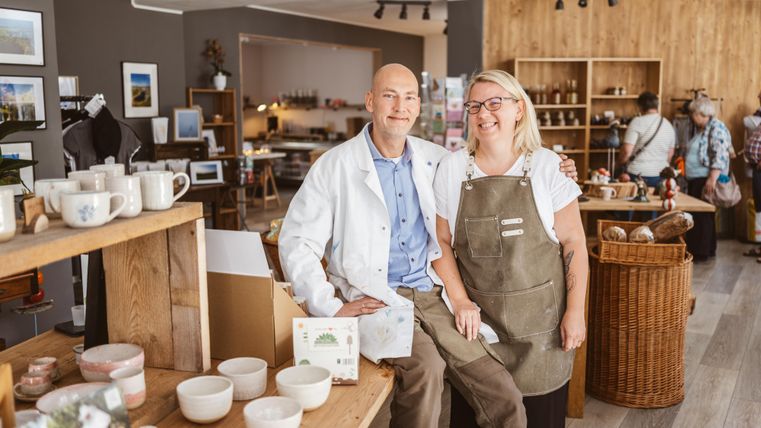 Un homme en blouse blanche et une femme en tablier se tiennent côte à côte en souriant dans un magasin. Le magasin est rempli de céramiques et d'autres marchandises.