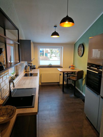 Eine moderne Küche mit Holzoberflächen und eleganten Geräten. Neben dem Fenster steht ein kleiner Tisch mit Stuhl und frischem Obst.