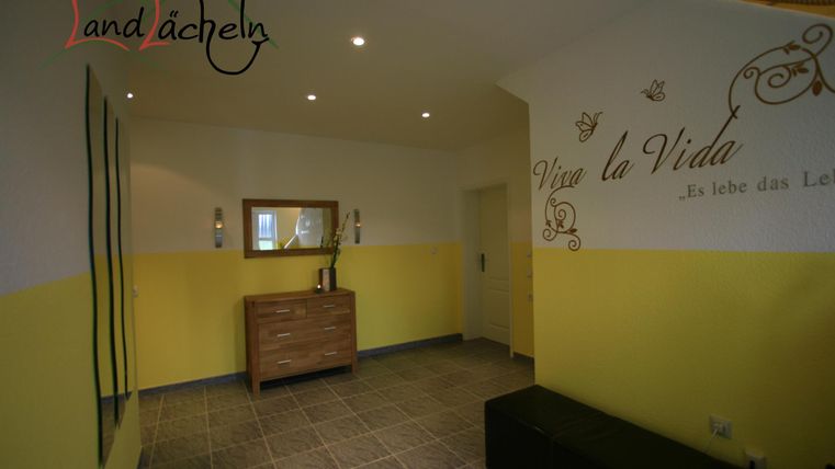 Ein heller Flur mit gelben Wänden und grauen Fliesen. Links steht eine moderne Kommode und eine Wand mit einem inspirierenden Spruch.