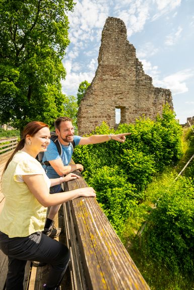 Ein mann und eine Frau lehnen an das Geländer einer Brücke, die zu einer Burgruine führt. Um die Burg und die Brücke herum stehen dicht bewachsene Bäume.