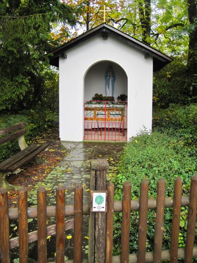 Eine kleine Kapelle mit einer Marienstatue, umgeben von Bäumen und einem Holzzaun.