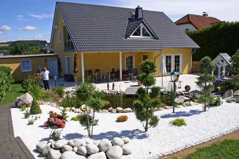 Een mooi huis met een gele gevel en een grijs dak. De tuin is verzorgd met stenen, planten en een kleine vijver.