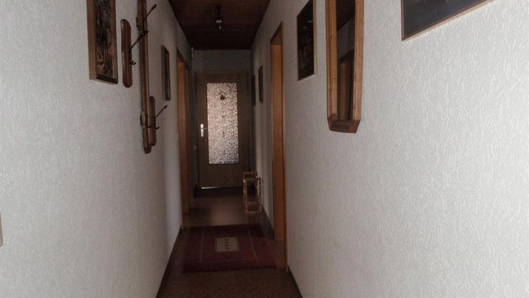 Ein heller Flur mit weißen Wänden und einem Holzdecken. An den Wänden hängen Bilder und ein Spiegel, am Boden liegt ein Teppich.