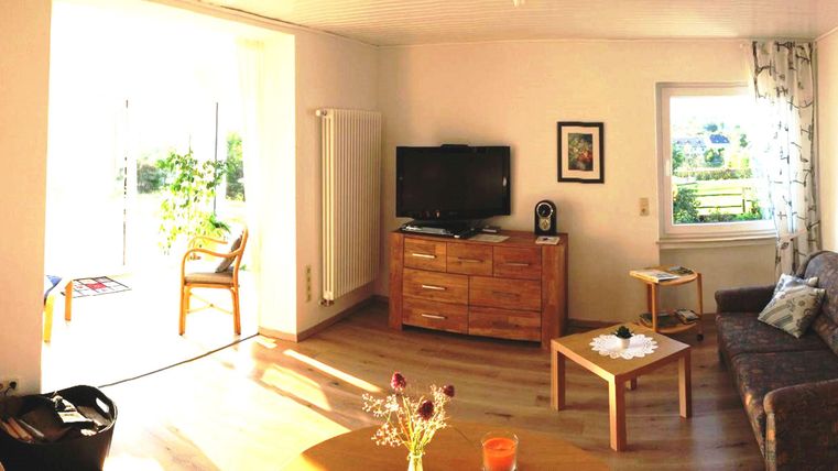 Ein helles Wohnzimmer mit einem Sofa, einem Tisch und einem Fernseher. Durch ein Fenster blickt man auf einen schönen Außenbereich.