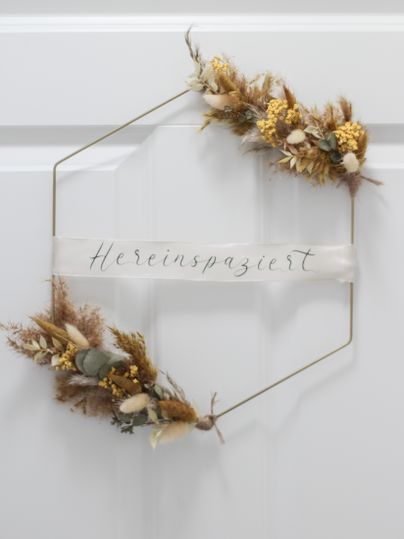 Ein dekorativer Türkranz in Form eines Hexagons. Mit getrockneten Blumen und einer eleganten Schriftrolle, die "Hereinspaziert" sagt.
