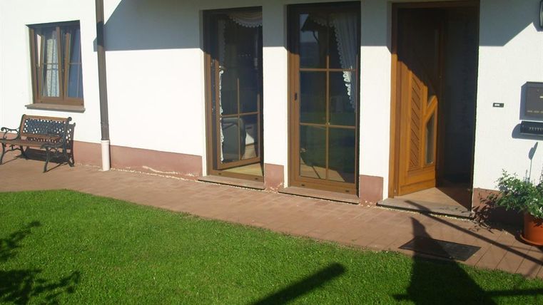 Ein gepflegter Garten mit grünem Rasen und zwei Eingängen zu einem Gebäude. Eine Bank lädt zum Verweilen ein.