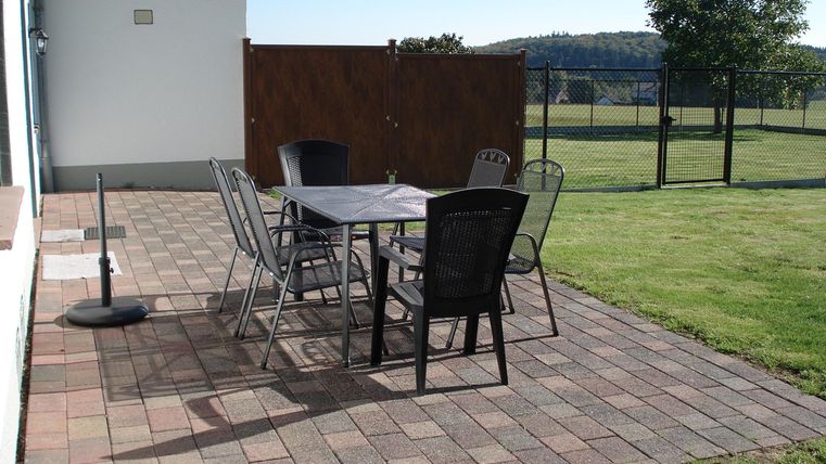 Een gezellige terras met een tafel en stoelen. Op de achtergrond zijn een grasveld en een hek te zien.