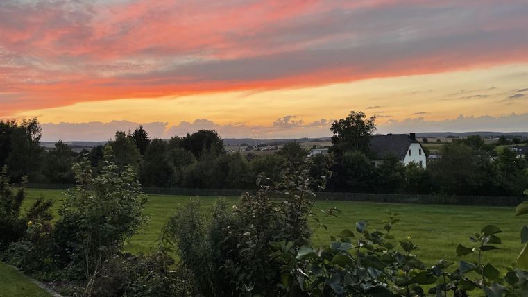 Ein wunderschöner Sonnenuntergang mit bunten Wolken über grünen Feldern. Im Hintergrund sind Häuser und Bäume zu sehen.