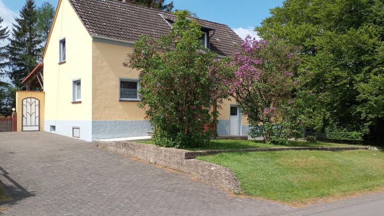 Ein gelbes Haus umgeben von Bäumen und Sträuchern. Der gepflasterte Weg führt zur Einfahrt.