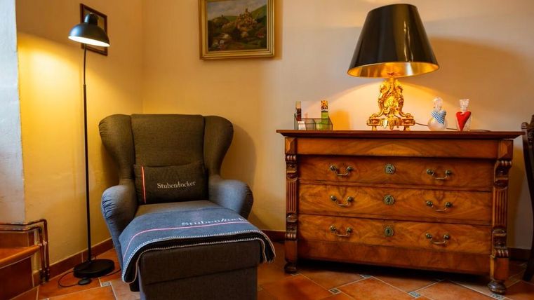 Ein gemütlicher Sessel steht neben einer Lampe in einem warmen Raum. Ein antiker Schrank mit dekorativen Details ergänzt die Einrichtung.