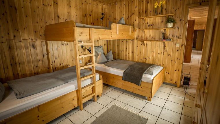 Ein einfaches Zimmer mit zwei Betten, einem Hochbett und einem Einzelbett aus Holz. Die Wände und der Boden sind ebenfalls aus Holz, was eine gemütliche Atmosphäre schafft.