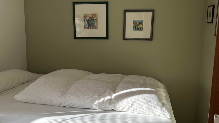 Ein gemütliches Schlafzimmer mit einem hohen Bett und frischen Bettdecken. An der Wand hängen zwei Bilder, und das Licht fällt sanft durch ein Fenster.