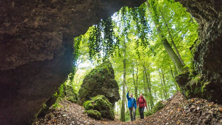 Zwei Personen wandern auf einen Höhleneingang aus Stein zu und zeigen auf die Felsen runderhum. Der Wald vor den Höhlen ist voller grün blühender hoher Bäume.