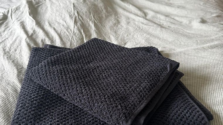 Ein Bett mit einem zerknitterten Bettlaken und zwei Kissen. Darauf liegen zwei faltete Handtücher in dunklem Farbton.