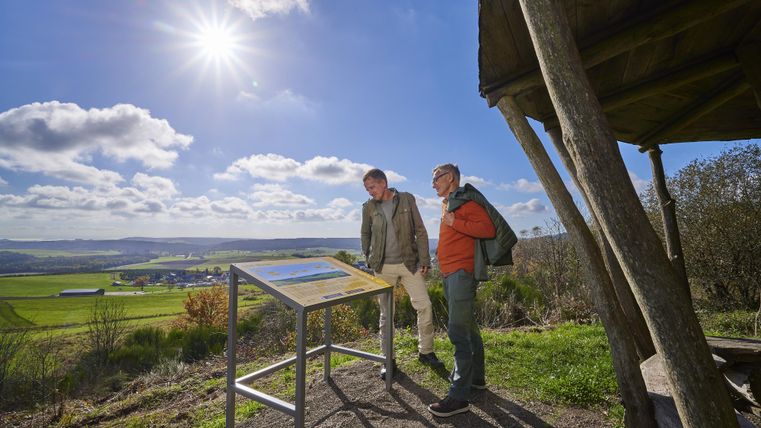 Deux personnes se tiennent devant un panneau d'information dans la nature. À l'arrière-plan, on voit un paysage verdoyant sous un ciel dégagé.