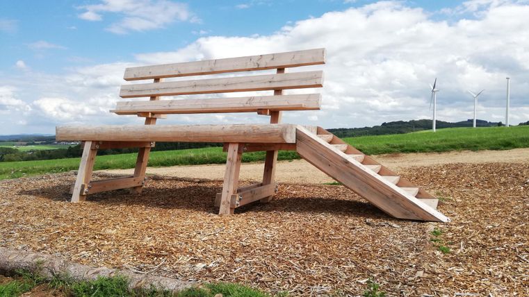 Eine übergroße Holzbank mit Treppe steht auf einem Feld. Im Hintergrund sind Windräder und ein bewölkter Himmel zu sehen.