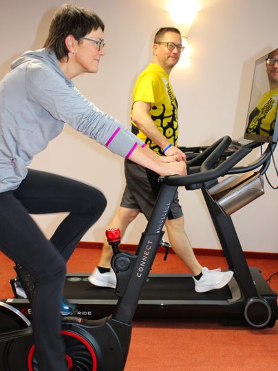 Deux personnes s'entraînent dans une salle de sport. Une femme fait du vélo d'appartement pendant qu'un homme court sur un tapis de course.