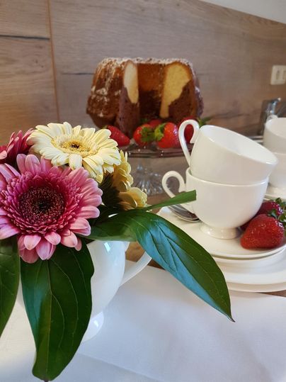 Ein hübsches Arrangement aus Blumen in einer Teekanne und weißen Tassen. Im Hintergrund sieht man einen Kuchen und frische Erdbeeren.