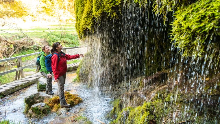 Zwei Personen betrachten den moosbedeckten Wasserfall in der grünen Waldlandschaft. Ein Holzsteg führt über den Bach.