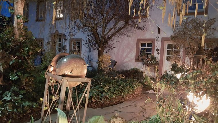 Een charmant huis omringd door een mooie tuin. Op de voorgrond staat een interessante sculptuur en het landschap is bij schemering verlicht.