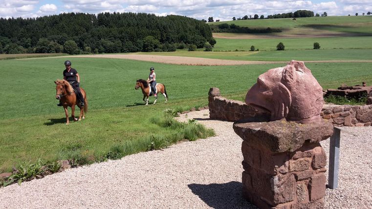 Twee ruiters op kleine paarden rijden door een groene landschap. Op de voorgrond staat een stenen object.
