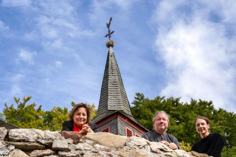 Trois personnes se tiennent devant un mur en pierre avec un clocher en arrière-plan. Le ciel est bleu avec quelques nuages.