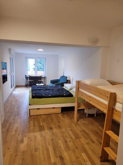 Une chambre simple avec un sol en bois, deux lits et une table. De grandes fenêtres offrent beaucoup de lumière.