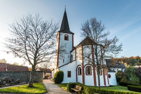 Kirche mit Kirchturm  in weiß gehalten mit ziegelsteinroten Akzenten, im Vordergrund Wiese mit zwei Bäumen, gutes Wetter