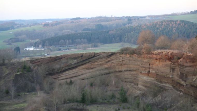 Eine beeindruckende Klippe mit roten Felsen und bewaldeten Hügeln im Hintergrund. Der Himmel ist klar und die Landschaft wirkt ruhig und unberührt.