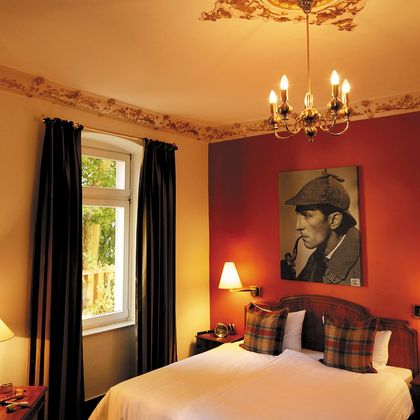 Une chambre confortable avec un grand lit et un élégant lustre. Un portrait de Sherlock Holmes est accroché sur un mur.