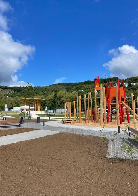 Een nieuwe houten speeltuin met rode elementen staat op een bestraatte vlak. Op de achtergrond zijn er bomen en een beboste heuvel te zien.