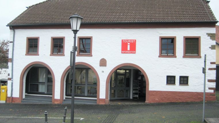 Ein einfaches, zweigeschossiges Gebäude mit einem roten Schild und großen Fenstern. Die Fassade ist weiß mit braunen Akzenten und einem traditionellen Satteldach.