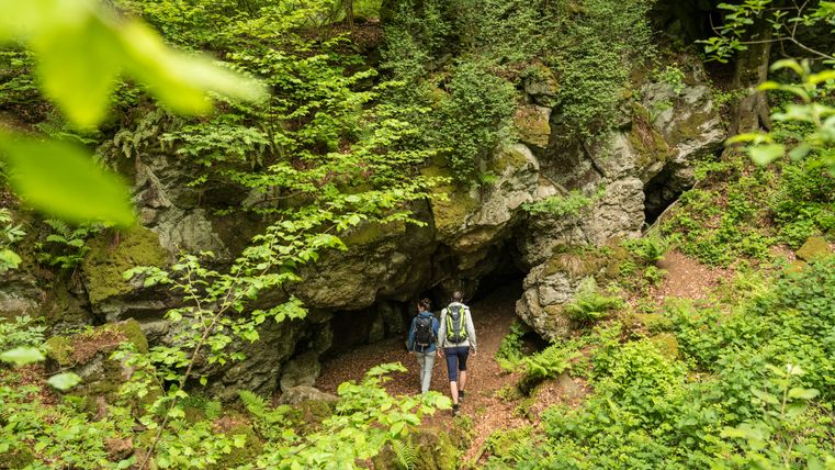 Two hikers surrounded by rocks overgrown with plants. They are walking towards the entrance to a cave.