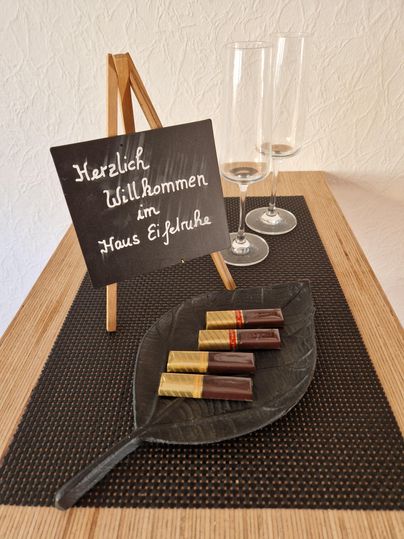 A welcome sign stands on a table, surrounded by champagne glasses. On a sheet, chocolate bars are laid out in various colors.